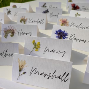 May include: White place cards with pressed flowers and handwritten names, including "Kathie", "Henry", "Nancy", "Marshall", "Darren", "Melissa", "Brad", "Kendall", "Jake", "Dana", "Tanke", and "Maria".