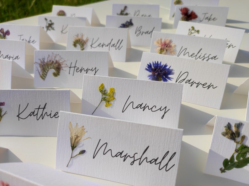 May include: White place cards with pressed flowers and handwritten names, including "Kathie", "Henry", "Nancy", "Marshall", "Darren", "Melissa", "Brad", "Kendall", "Jake", "Dana", "Tanke", and "Maria".