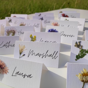 May include: White place cards with pressed flowers and names written in black ink. The names include Kathie, Marshall, Laura, Darren, and others.