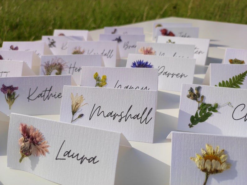 May include: White place cards with pressed flowers and names written in black ink. The names include Kathie, Marshall, Laura, Darren, and others.