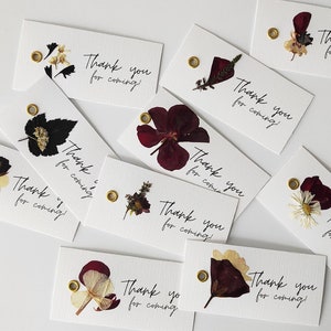 May include: A collection of white thank you tags with pressed flowers and gold eyelets. The tags have the text "Thank you for coming!" written in black calligraphy.