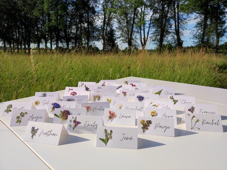 May include: A set of white place cards with pressed flowers and handwritten names, such as "Angie", "Nathan", "Melis", "Laura", "Jen&eacute;", "Paige", "Anna", "Julie", "Chris", "Frances", and "Rachel".