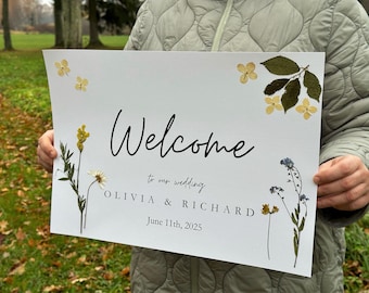 May include: A white welcome sign with pressed flowers and greenery. The sign reads "Welcome to our wedding Olivia & Richard June 11th, 2025".