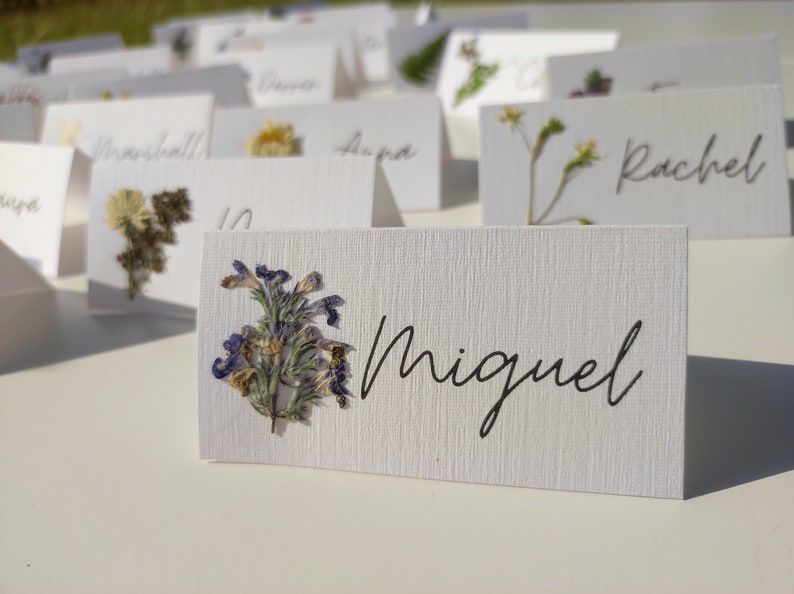 May include: White place cards with pressed flowers and the names "Miguel", "Rachel", "Manchella", "Dana", "Laura", and "Tanya" written in black ink.
