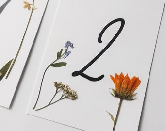 May include: A white card with the number "2" written in black cursive. The card is decorated with pressed flowers, including a small blue flower, a cluster of white flowers, and a large orange flower.