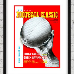 May include: A red and white poster advertising the 8th Annual Football Classic between the Philadelphia Eagles and the Green Bay Packers on September 13, 1945 at Municipal Stadium. The poster features a close-up of a football being held by a hand.