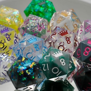 May include: A collection of polyhedral dice in various colors, including clear, green, yellow, and gray. Each die features a crown symbol and numbers. Some dice have glitter or colored accents. The dice are designed for tabletop gaming.
