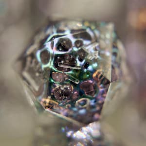 May include: Close-up of a faceted, clear crystal with internal inclusions. The crystal's interior features a complex arrangement of dark, circular and linear elements, with iridescent colors of green, purple, and gold.