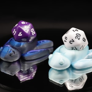May include: Two 3D-printed turtle figurines, one purple and blue, the other light blue, each supporting a twenty-sided die. The dice are white with black numbers and purple with white numbers. The figurines are on a reflective black surface.