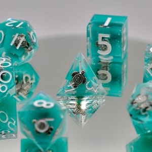 May include: Set of teal and silver dice with a turtle design. The dice are made of clear plastic with a teal tint and have silver turtles embedded in them. The numbers on the dice are white.