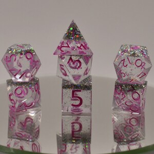 May include: Set of five clear dice with pink numbers and glitter embedded in the plastic. The dice are in a pyramid shape with the top die being a d4, the next die being a d6, and the bottom die being a d20. The dice are on a reflective surface.