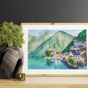 Puede incluir: Pintura a la acuarela de un pueblo junto al lago con un campanario, enmarcada en madera clara. La obra representa un paisaje sereno con monta&ntilde;as verdes, agua azul y edificios coloridos. Un jarr&oacute;n decorativo y una vela est&aacute;n en primer plano.