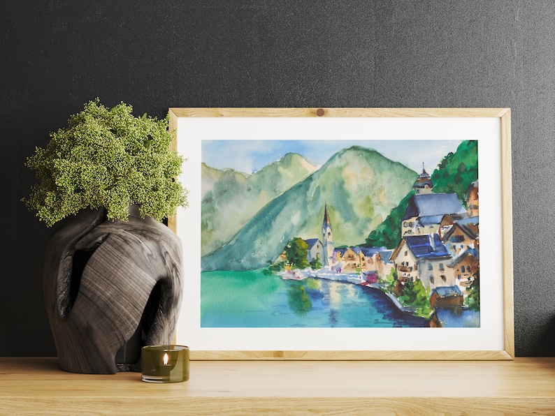 Puede incluir: Pintura a la acuarela de un pueblo junto al lago con un campanario, enmarcada en madera clara. La obra representa un paisaje sereno con monta&ntilde;as verdes, agua azul y edificios coloridos. Un jarr&oacute;n decorativo y una vela est&aacute;n en primer plano.