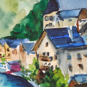Puede incluir: Pintura a la acuarela de un pueblo europeo enclavado contra una monta&ntilde;a verde. Los edificios tienen paredes marrones y beige con tejados azules. La escena incluye un r&iacute;o en primer plano, con follaje verde y morado.