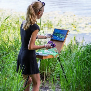 May include: A woman in a black dress paints outdoors by a lake. She is using a wooden easel and a palette knife to apply paint to a canvas. The canvas depicts a landscape with water and greenery.