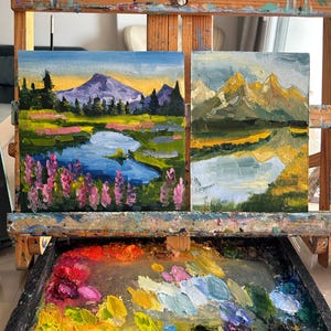May include: Two small oil landscape paintings displayed on a wooden easel. The left painting shows a purple mountain, a blue lake, and pink wildflowers. The right painting depicts golden mountains and a river. A paint palette with colorful oil paint sits below.
