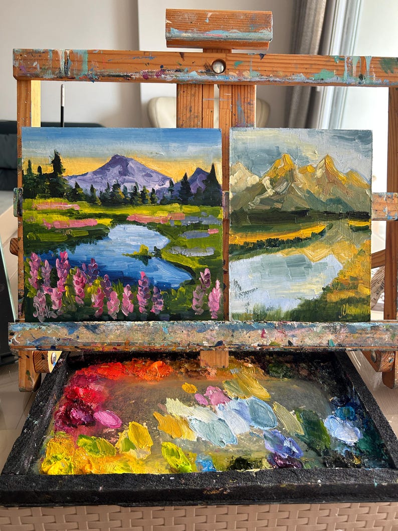 May include: Two small oil landscape paintings displayed on a wooden easel. The left painting shows a purple mountain, a blue lake, and pink wildflowers. The right painting depicts golden mountains and a river. A paint palette with colorful oil paint sits below.