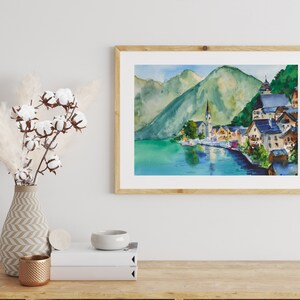 Puede incluir: Pintura a la acuarela de un pueblo junto al lago con una iglesia y monta&ntilde;as al fondo. La obra de arte est&aacute; enmarcada en un marco de madera clara y se exhibe sobre una superficie de madera. Un jarr&oacute;n con hierba de la pampa y tallos de algod&oacute;n est&aacute; a la izquierda.