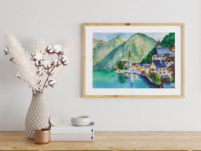 Puede incluir: Pintura a la acuarela de un pueblo junto al lago con una iglesia y monta&ntilde;as al fondo. La obra de arte est&aacute; enmarcada en un marco de madera clara y se exhibe sobre una superficie de madera. Un jarr&oacute;n con hierba de la pampa y tallos de algod&oacute;n est&aacute; a la izquierda.