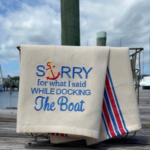 May include: A white tea towel with blue embroidery that reads "SORRY for what I said WHILE DOCKING The Boat" with a red and yellow anchor above the word "SORRY". The tea towel is folded and hanging from a metal rack on a wooden dock.