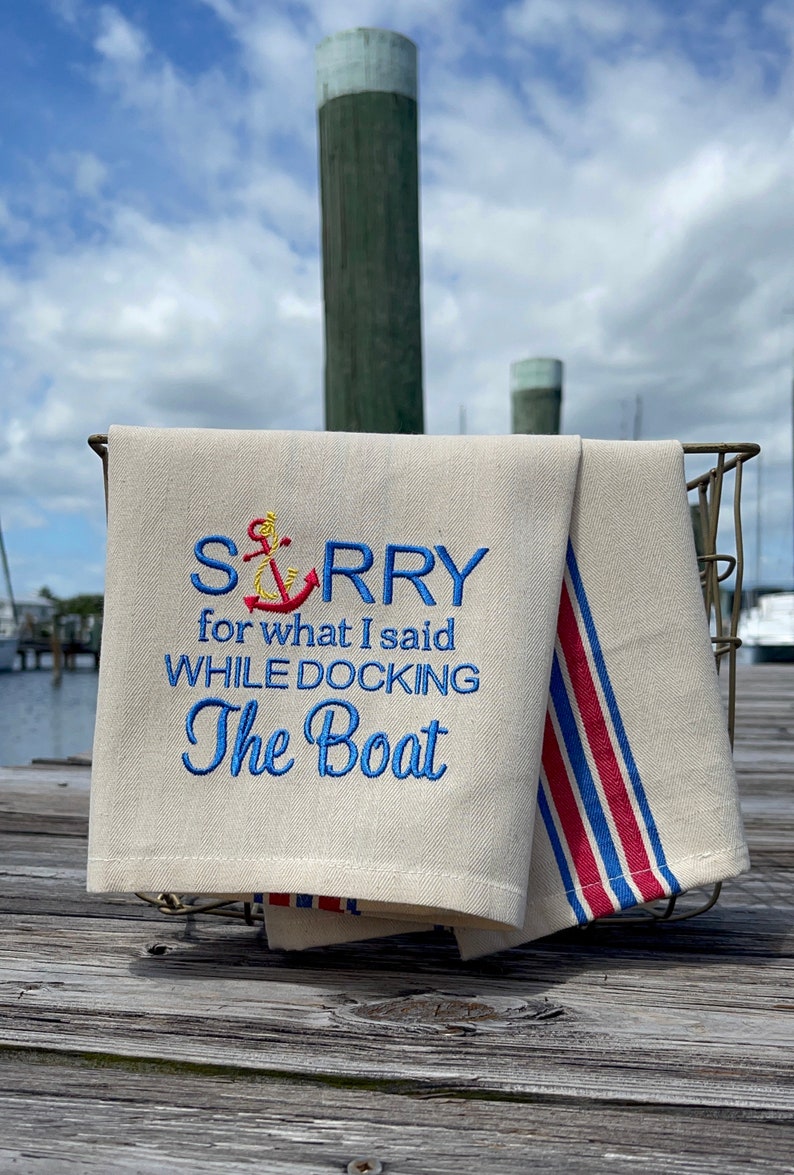 May include: A white tea towel with blue embroidery that reads "SORRY for what I said WHILE DOCKING The Boat" with a red and yellow anchor above the word "SORRY". The tea towel is folded and hanging from a metal rack on a wooden dock.