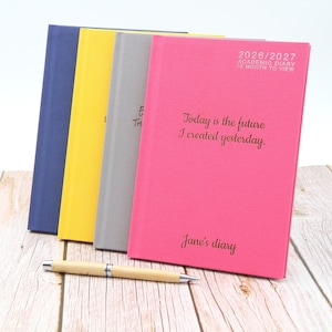 Peut inclure: Quatre agendas académiques à couverture rigide, bleus, jaunes, gris et roses, avec un stylo en bois. L'agenda rose porte le texte "2026/2027 Academic Diary 18 Month to View" et "Today is the future I created yesterday. Jane's diary."