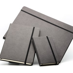May include: Three gray notebooks with black elastic closures. The notebooks are stacked on top of each other, with the largest notebook on the bottom and the smallest notebook on top.