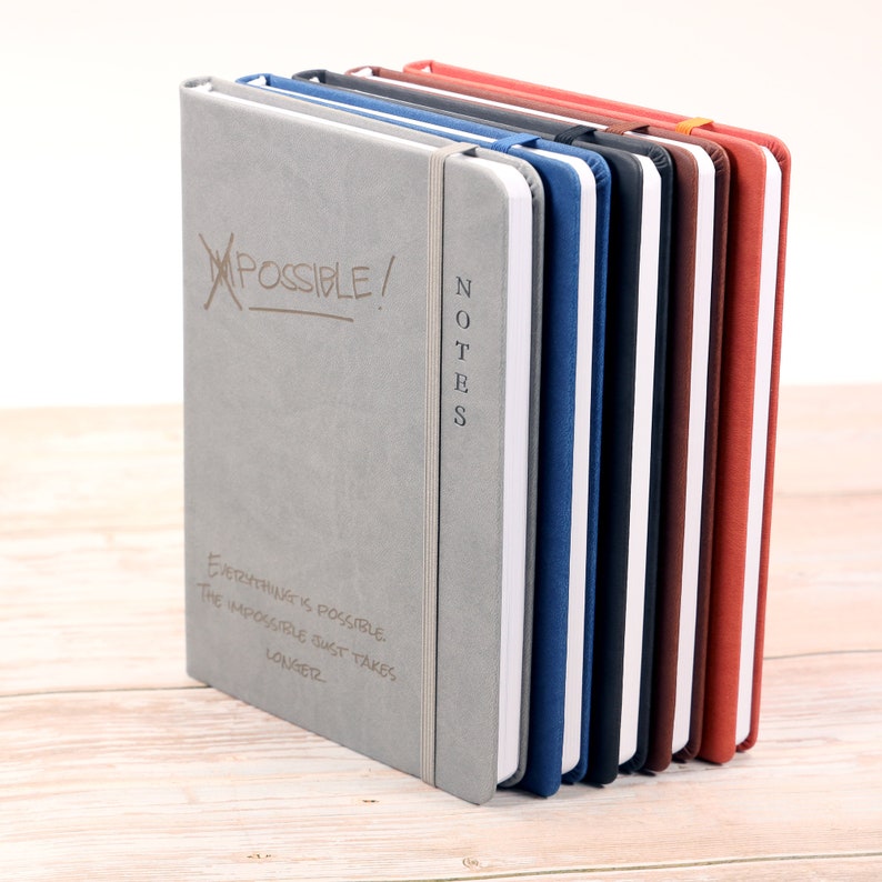May include: A stack of eight gray, blue, black, white, brown, and red notebooks with the word "NOTES" written vertically on the front of the gray notebook. The front of the gray notebook also has the words "IMPOSSIBLE!" crossed out and the text "Everything is possible. The impossible just takes longer." written below.