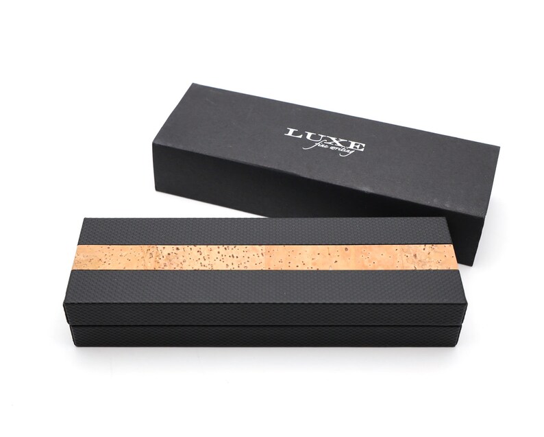 May include: Two rectangular boxes for writing instruments. One box is solid black with the white text LUXE fine writing. The other box features a textured black finish with a horizontal strip of natural tan cork running across the center.