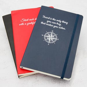 May include: Three notebooks in black, red, and navy blue. The navy blue notebook features the text "Travel is the only thing you can buy, that makes you richer" and a compass rose graphic. The red notebook has the text "Start each day with a grateful".