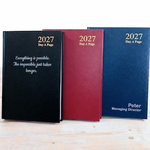Op de afbeelding: Drie 2027 Day A Page planners in zwart, bordeauxrood en marineblauw. De zwarte planner bevat het citaat Everything is possible. The impossible just takes longer. De marineblauwe planner is gepersonaliseerd met de tekst Peter Managing Director.