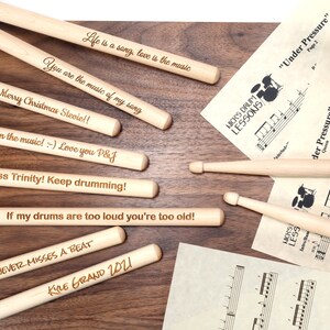 May include: A set of wooden drumsticks with personalized messages engraved on them. The messages include "Life is a song, love is the music", "You are the music of my song", "Merry Christmas Stevie!!", "Lose yourself in the music! :-) Love you P&J", "To Jamie from Miss Trinity! Keep drumming!", "If my drums are too loud you're too old!", "THE MAN WHO NEVER MISSES A BEAT", and "KIRE GRAND 2021".