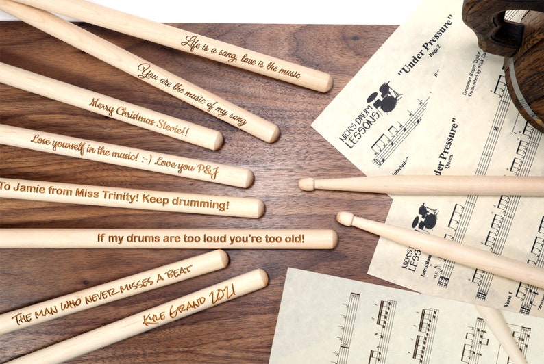 May include: A set of wooden drumsticks with personalized messages engraved on them. The messages include "Life is a song, love is the music", "You are the music of my song", "Merry Christmas Stevie!!", "Lose yourself in the music! :-) Love you P&J", "To Jamie from Miss Trinity! Keep drumming!", "If my drums are too loud you're too old!", "THE MAN WHO NEVER MISSES A BEAT", and "KIRE GRAND 2021".