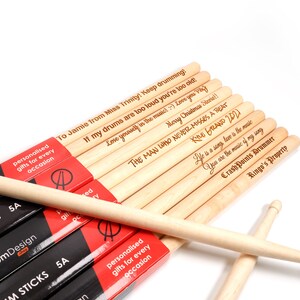 May include: A set of wooden drum sticks with personalized messages engraved on them. The messages include "To Jamie from Miss Trinity! Keep drumming! If my drums are too loud you're too old! Love yourself in the music! Love you Vay! Merry Christmas Steve!! The Man Who Never Misses A Beat!  Nine Grand 2021! Life is a song love is the music You are the music of my song Crash Points Drummer Ringo's Property". The sticks are packaged in black and red packaging that says "Custom Design Drum Sticks 5A".