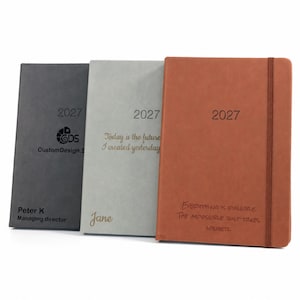Può includere: Tre agende rettangolari in grigio antracite, grigio chiaro e terracotta, ciascuna contrassegnata con 2027. L'agenda antracite presenta un logo e Peter K Managing director. Quella grigia riporta Today is the future I created yesterday e Jane. Quella terracotta riporta Everything is possible. The impossible just takes longetz.