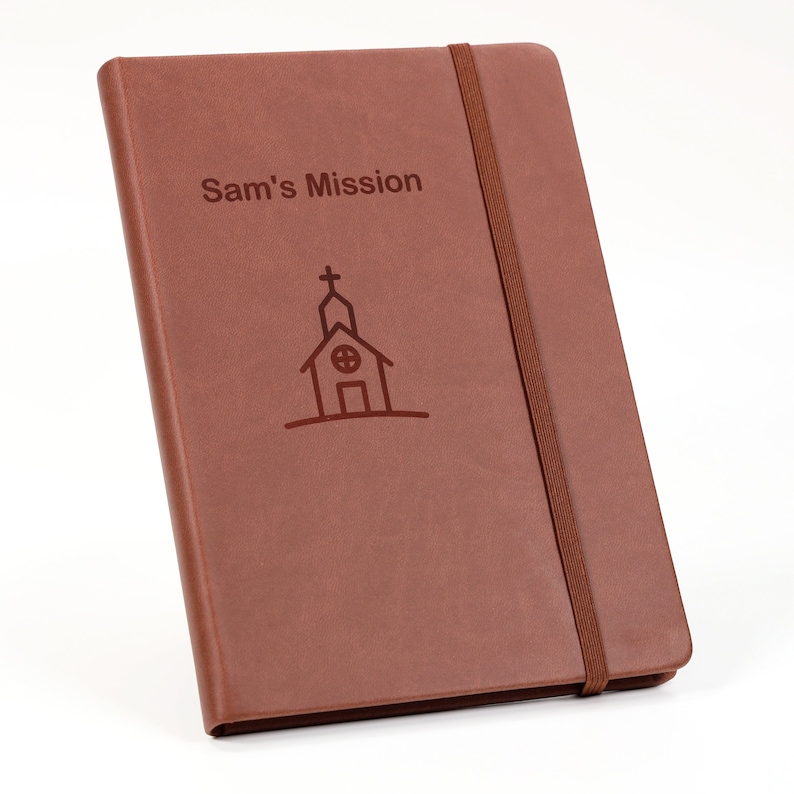 May include: A brown leather journal with a black elastic closure. The front cover has the text "Sam's Mission" and a simple line drawing of a church.
