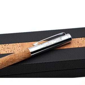 May include: A ballpoint pen with a natural cork barrel and a polished silver-tone metal cap featuring a pocket clip. The cap is engraved with the name Saphia Scott in a cursive script font. The pen rests on a black and cork textured case.