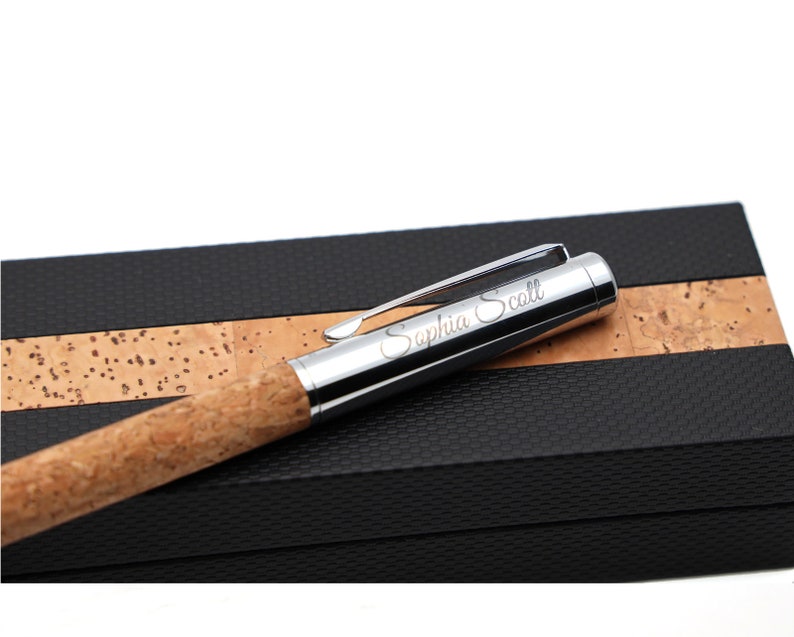 May include: A ballpoint pen with a natural cork barrel and a polished silver-tone metal cap featuring a pocket clip. The cap is engraved with the name Saphia Scott in a cursive script font. The pen rests on a black and cork textured case.