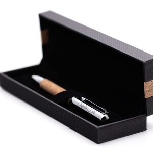 May include: A ballpoint pen with a light brown cork barrel and a polished silver cap engraved with the name Sophia Scott. The pen rests inside a rectangular black presentation box with a textured exterior and a cork accent on the side.