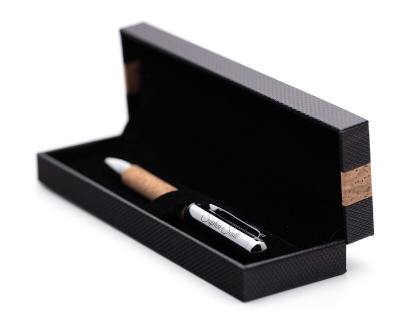 May include: A ballpoint pen with a light brown cork barrel and a polished silver cap engraved with the name Sophia Scott. The pen rests inside a rectangular black presentation box with a textured exterior and a cork accent on the side.