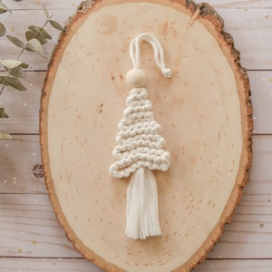Può includere: Un ornamento per albero di Natale in macramé bianco con un perno di legno in cima e un lungo fiocco. L'ornamento è esposto su una fetta di legno.