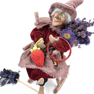 以下が含まれることがあります： 木製のロッキングチェアに座った、気まぐれな魔女の人形。バーガンディ色のベルベットのドレスと、お揃いの帽子を着用しています。赤いフープと紫の花のブーケを持っています。かぎ針編みのイチゴとほうきがシーンを完成させています。