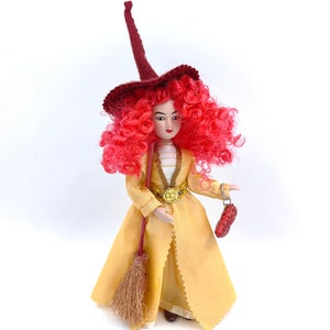 Può includere: Una bambola con i capelli rossi che indossa un vestito giallo e un cappello da strega rosso. Tiene una scopa e una piccola borsa rossa.