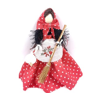 Puede incluir: Una muñeca hecha a mano con cabello negro, un vestido de lunares rojos y blancos, un delantal blanco con flores bordadas y un pañuelo rojo y blanco de lunares. La muñeca sostiene una escoba y un bulbo de ajo blanco.