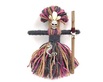 Op de afbeelding: Een handgemaakte garenpop met een schedelhoofd, roze en bruin garenhaar en een stok met een schedelbedel. De pop draagt een rok van roze, bruin en grijs garen en heeft een zilveren zeepaardje en sleutelbedel aan haar taille.