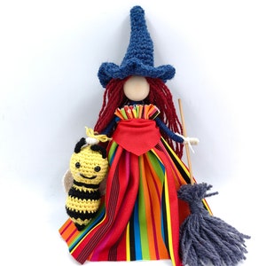 May include: A handmade witch doll with a blue hat, red yarn hair, and a colorful striped dress. She is holding a crocheted bee and a broom.
