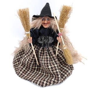 Op de afbeelding: Een heksenpop met lang blond haar, die een zwarte jurk draagt met een bruin-wit geruit patroon en een zwarte heksenhoed. Ze houdt twee bezems in haar handen.