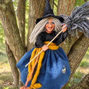 May include: A witch doll featuring a black pointed hat, long gray yarn hair, and a painted face with blue eyes. The doll wears a black long-sleeved top, a mustard yellow sash, and a blue skirt with a small silver cat charm. It holds a broom.