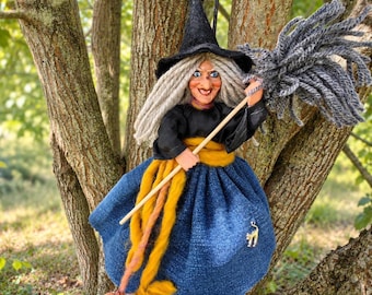 Può includere: Una bambola strega con cappello a punta nero, lunghi capelli di lana grigia e un volto dipinto con occhi azzurri. La bambola indossa una maglia nera a maniche lunghe, una fascia giallo senape e una gonna blu con un piccolo ciondolo a gatto.