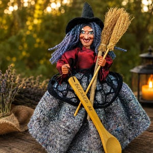 Può includere: Una bambola strega con capelli blu, un cappello a punta nero e un top rosso scuro. La bambola indossa una gonna grigia testurizzata e tiene in mano una piccola scopa di paglia e un cucchiaio di legno con la scritta BLESSED BE incisa sul manico.